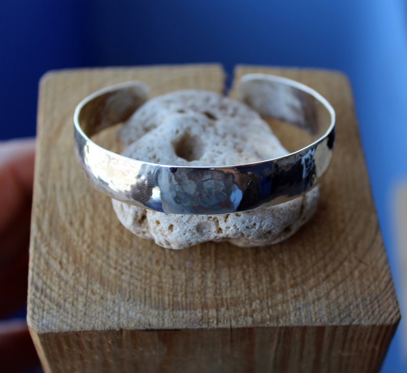 May include: A silver hammered cuff bracelet with a smooth, rounded surface. The bracelet is displayed on a piece of light brown wood.