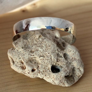 May include: A silver hammered bangle bracelet with a smooth, rounded surface. The bracelet is resting on a piece of rough, textured rock.
