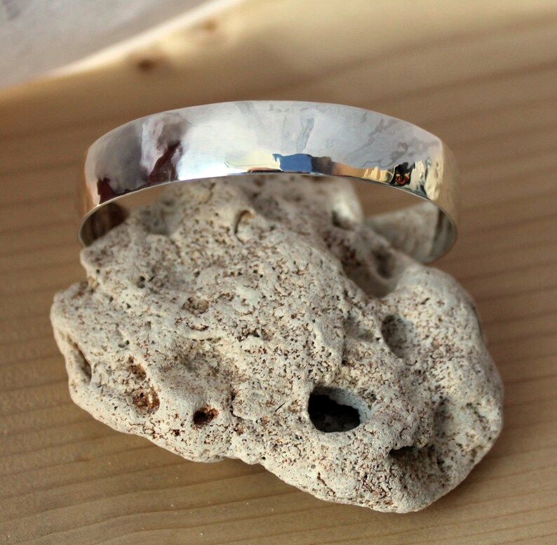 May include: A silver hammered bangle bracelet with a smooth, rounded surface. The bracelet is resting on a piece of rough, textured rock.