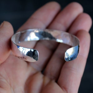 May include: A silver hammered cuff bracelet with a smooth, polished finish. The bracelet is open ended and has a slightly curved shape.