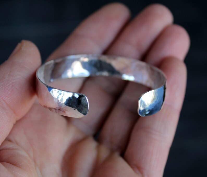 May include: A silver hammered cuff bracelet with a smooth, polished finish. The bracelet is open ended and has a slightly curved shape.