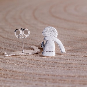 May include: A pair of silver-colored stud earrings. One earring is a post with a butterfly backing, and the other is a small figure wearing a scarf. The earrings are on a light brown wooden surface.