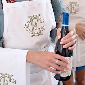 May include: A white apron featuring an embroidered olive green monogram with the letters E and C intertwined. The design is displayed on the chest and on the lower front pocket area. The apron has white fabric ties at the waist and neck.