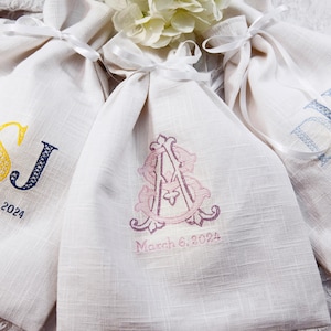 K&ouml;nnte beinhalten: Drei cremefarbene Leinen-Kordelzugbeutel, jeweils mit Initialen und Datum bestickt. Einer zeigt die Initialen "GSJ" in Marineblau und Gelb und das Datum "14. September 2024". Ein anderer hat rosa Initialen "A" und "M" mit "6. M&auml;rz 2024". Der dritte zeigt blaue Initialen "DK" und "8. Juni 2024."