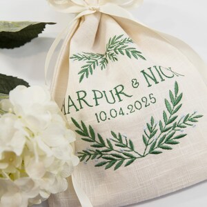 K&ouml;nnte beinhalten: Ein beiger Leinenbeutel mit gr&uuml;n gesticktem Text und einem Lorbeerkranz-Design. Der Text lautet "MARPUR & NICK 10.04.2025". Eine wei&szlig;e Blume und Gr&uuml;nzeug befinden sich im Hintergrund.