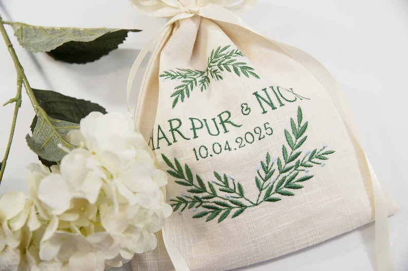 K&ouml;nnte beinhalten: Ein beiger Leinenbeutel mit gr&uuml;n gesticktem Text und einem Lorbeerkranz-Design. Der Text lautet "MARPUR & NICK 10.04.2025". Eine wei&szlig;e Blume und Gr&uuml;nzeug befinden sich im Hintergrund.