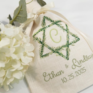 K&ouml;nnte beinhalten: Beiger Leinen-Geschenkbeutel mit gr&uuml;n gesticktem Sterndesign und dem Buchstaben "C" in der Mitte. Der Beutel ist mit einem cremefarbenen Band gebunden und tr&auml;gt die Namen "Ethan & Lindsay" und das Datum "10.25.2025". Eine wei&szlig;e Hortensienbl&uuml;te ist im Vordergrund.
