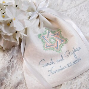 K&ouml;nnte beinhalten: Ein cremefarbener Beutel mit Kordelzug und einem Davidstern-Design in Rosa, Blau und Gr&uuml;n. Der Beutel ist mit den Namen "Sarah und Stephen" und dem Datum "28. November 2024" bestickt. Wei&szlig;e Blumen und Schleife im Hintergrund.