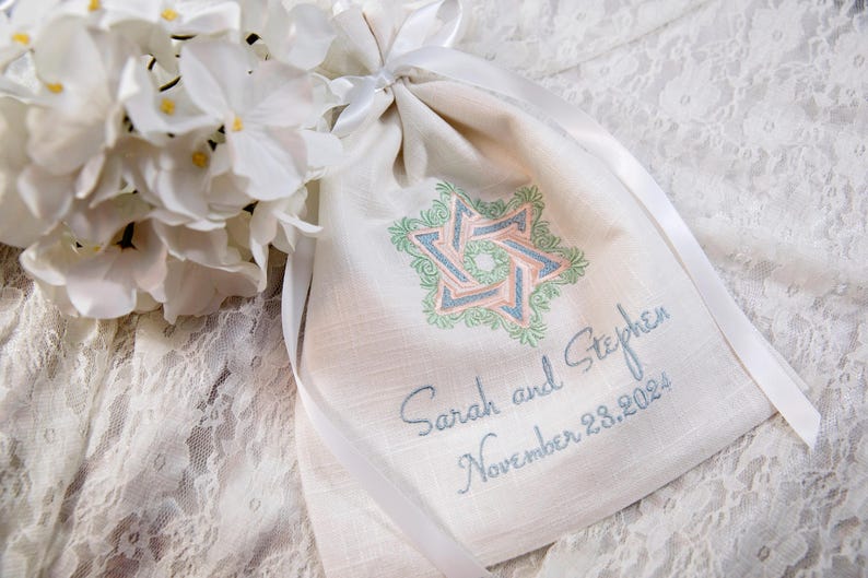 K&ouml;nnte beinhalten: Ein cremefarbener Beutel mit Kordelzug und einem Davidstern-Design in Rosa, Blau und Gr&uuml;n. Der Beutel ist mit den Namen "Sarah und Stephen" und dem Datum "28. November 2024" bestickt. Wei&szlig;e Blumen und Schleife im Hintergrund.