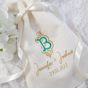 K&ouml;nnte beinhalten: Cremefarbener Leinen-Geschenkbeutel mit einem gr&uuml;n-goldenen gestickten Initialen, den Namen "Jennifer & Joshua" und dem Datum "09.03.2023". Der Beutel ist mit einem wei&szlig;en Satinband gebunden. Wei&szlig;e Blumen sind im Hintergrund.