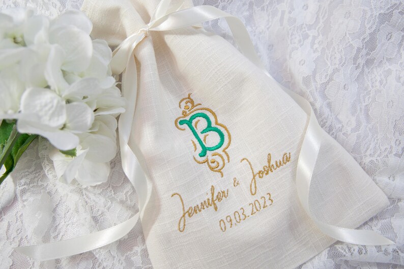 K&ouml;nnte beinhalten: Cremefarbener Leinen-Geschenkbeutel mit einem gr&uuml;n-goldenen gestickten Initialen, den Namen "Jennifer & Joshua" und dem Datum "09.03.2023". Der Beutel ist mit einem wei&szlig;en Satinband gebunden. Wei&szlig;e Blumen sind im Hintergrund.