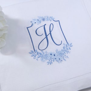 May include: White pillowcase with a blue embroidered floral design and the letter "H" in a shield shape. The pillowcase has a stitched border. A bouquet of white flowers is in the upper left corner.