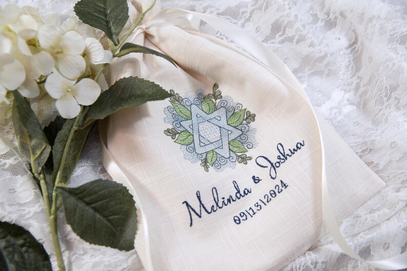 K&ouml;nnte beinhalten: Ein cremefarbener Kordelzugbeutel mit einem gestickten Davidstern-Design in Blau und Gr&uuml;n, umgeben von Bl&auml;ttern. Die Namen "Melinda & Joshua" und das Datum "09/13/2024" sind darunter gestickt. Eine wei&szlig;e Blume und Gr&uuml;nzeug sind im Hintergrund.