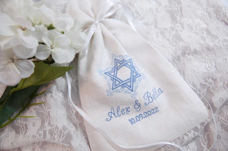 K&ouml;nnte beinhalten: Ein wei&szlig;er Leinenschnurbeutel mit blauem Davidstern-Design und den Namen "Alex & Bella" mit dem Datum "10.09.2022" in Blau gestickt. Wei&szlig;e Blumen und ein Band sind im Hintergrund.