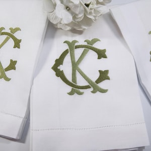May include: White linen hand towels with olive green embroidered monograms. The towels are folded and arranged, with a floral accent in the background. The monogram features the letters "KC" in an elegant, stylized font.