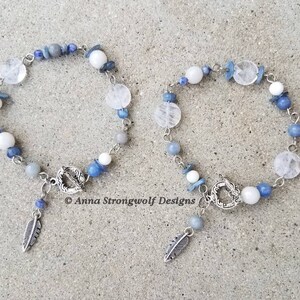 May include: Two beaded bracelets with a mix of white, blue, and clear gemstones. Each bracelet has a silver clasp and a dangling feather charm. The bracelets are displayed on a light gray surface.