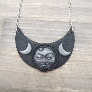 May include: A crescent moon pendant necklace with a silver chain. The black pendant features a central face with closed eyes, framed by a rope design, and two silver crescent moons. The necklace is a celestial-themed accessory.