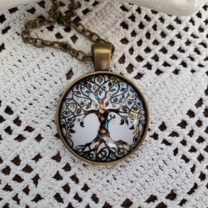 May include: A round pendant necklace with a bronze-colored chain. The pendant features a detailed tree of life design with colorful leaves against a white background. The necklace is set on a white, open-weave fabric.