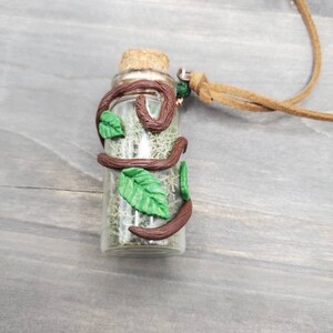 May include: A small glass bottle necklace filled with green moss and sealed with a cork. The bottle is adorned with brown clay vines and green leaves. A tan leather cord is attached for wearing.