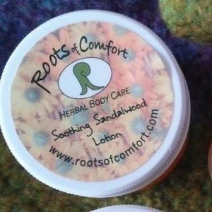 May include: White plastic jar with a label that reads "Roots of Comfort Herbal Body Care Soothing Sandalwood Lotion www.rootsofcomfort.com".