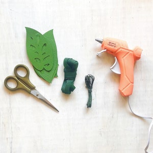 K&ouml;nnte beinhalten: Eine flache Anordnung von Bastelmaterialien f&uuml;r ein DIY-Projekt. Die Materialien umfassen gr&uuml;ne Papierbl&auml;tter, gr&uuml;nes Garn, eine Klebepistole und eine Schere. Die Klebepistole ist orange und hat den Text "Adhesive Hotmelt" auf der Seite.