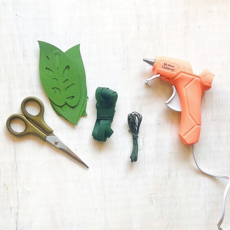K&ouml;nnte beinhalten: Eine flache Anordnung von Bastelmaterialien f&uuml;r ein DIY-Projekt. Die Materialien umfassen gr&uuml;ne Papierbl&auml;tter, gr&uuml;nes Garn, eine Klebepistole und eine Schere. Die Klebepistole ist orange und hat den Text "Adhesive Hotmelt" auf der Seite.
