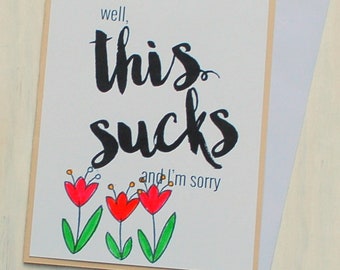 May include: A white card with black text that reads "well, this sucks and I'm sorry". There are three red flowers with green stems and yellow centers drawn on the card.