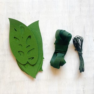 K&ouml;nnte beinhalten: Gr&uuml;ne Papierbl&auml;tter mit Monstera-Blattdesign, gr&uuml;nes Garn und eine Rolle gr&uuml;nes Band. Dies sind Bastelmaterialien f&uuml;r die Herstellung von Blumenarrangements.