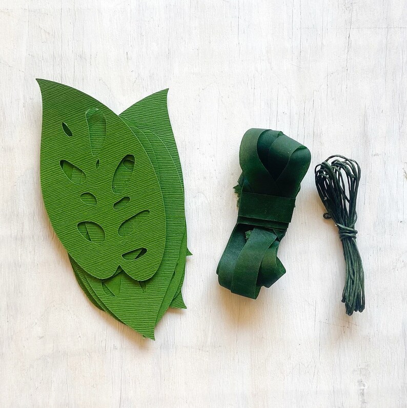 K&ouml;nnte beinhalten: Gr&uuml;ne Papierbl&auml;tter mit Monstera-Blattdesign, gr&uuml;nes Garn und eine Rolle gr&uuml;nes Band. Dies sind Bastelmaterialien f&uuml;r die Herstellung von Blumenarrangements.