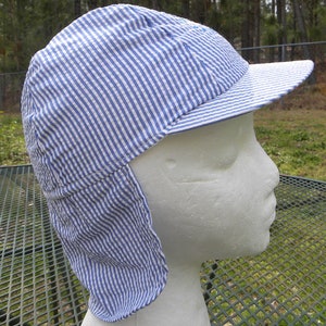 May include: A blue and white striped hat with a visor and ear flaps. The hat is made of a lightweight fabric and is designed to provide sun protection. The hat is displayed on a white mannequin head.