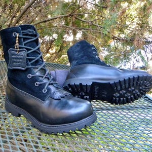 May include: A pair of black boots featuring a combination of smooth leather and suede textures. The boots have a lace-up front, a thick lugged rubber sole, and a small leather tag attached to the laces with the text Genuine Leather Upper.