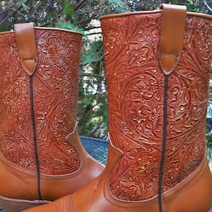 May include: A pair of brown leather cowboy boots with intricate floral tooling. The boots have a classic design with a pull strap and a pointed toe. The leather has a rich, warm tone, and the detailed carving adds a touch of elegance to the western-style footwear.