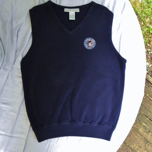 May include: Navy blue IZOD CLUB V-neck sweater vest. The vest has a small embroidered emblem on the chest and a tag at the neckline. The vest is made of a soft knit material. The vest is sleeveless.