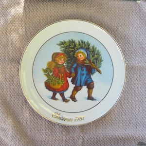 May include: A decorative plate with a gold rim and a central image of two children in winter attire carrying a Christmas tree and basket. The plate is white with a blue background and the text "Christmas 1981" in gold.