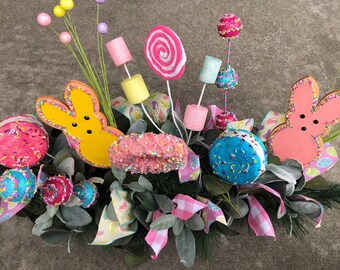 May include: A spring-themed centerpiece featuring a yellow and pink Peeps bunny, a pink donut, a blue and white cookie, a pink and white lollipop, and a variety of other candy decorations.
