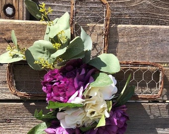 Op de afbeelding: Een rustiek draadkruis versierd met een boeket van paarse en witte bloemen en groen.