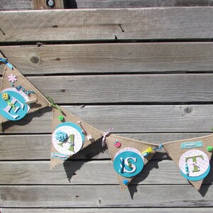 May include: A burlap banner with the word "Easter" spelled out in blue and white letters. Each letter is decorated with various embellishments, including flowers, buttons, and ribbons.
