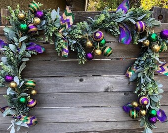 May include: A Mardi Gras garland with green, purple, and gold beads and ribbons. The garland is made of artificial greenery and is perfect for decorating for a Mardi Gras party.
