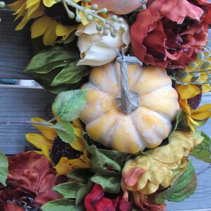 May include: A fall-themed wreath featuring sunflowers, pumpkins, and red and orange flowers. The wreath is made from artificial materials and is ideal for decorating a door or wall.