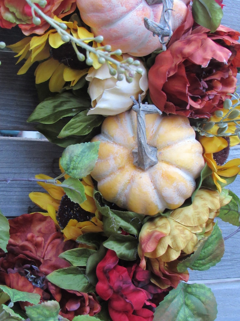 May include: A fall-themed wreath featuring sunflowers, pumpkins, and red and orange flowers. The wreath is made from artificial materials and is ideal for decorating a door or wall.