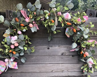 May include: A green eucalyptus garland with pink and white bunny decorations, pastel colored eggs, and orange carrots. The garland is hanging on a wooden fence.