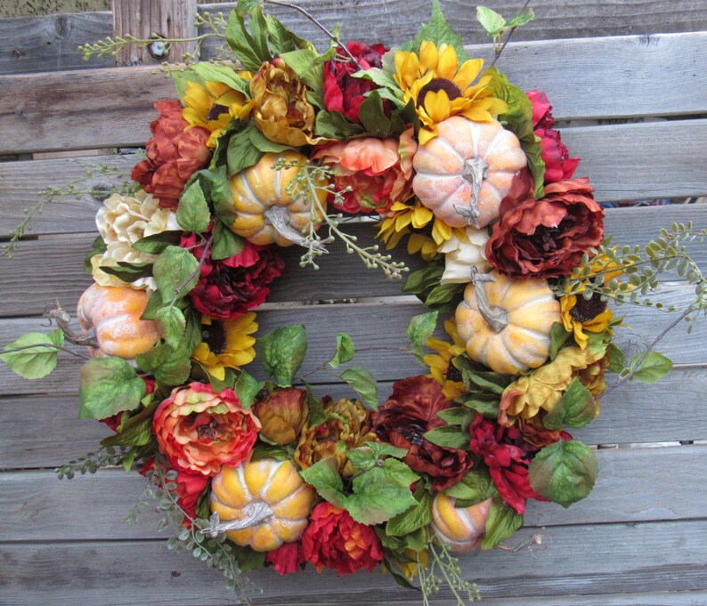 May include: A fall-themed wreath with red, orange, and yellow flowers, green leaves, and small pumpkins.