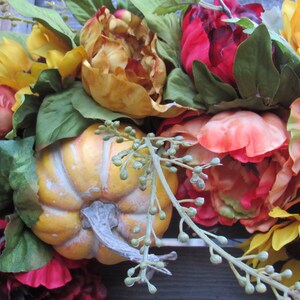 May include: A fall-themed floral arrangement featuring sunflowers, red and orange peonies, and small pumpkins. The arrangement is accented with green leaves and small green buds.