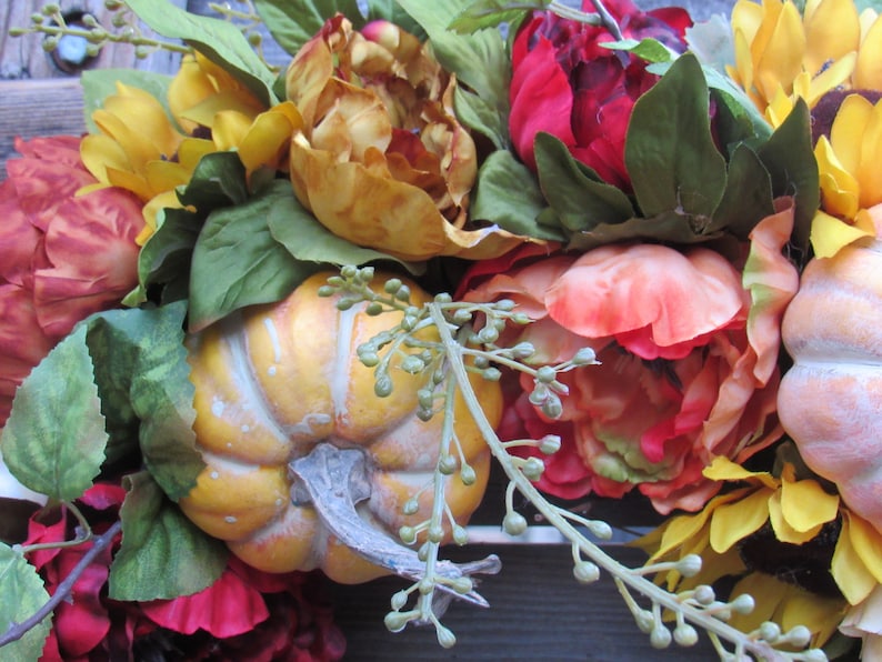 May include: A fall-themed floral arrangement featuring sunflowers, red and orange peonies, and small pumpkins. The arrangement is accented with green leaves and small green buds.