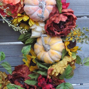 May include: A fall-themed wreath featuring red, orange, and yellow flowers, green leaves, and two faux pumpkins.