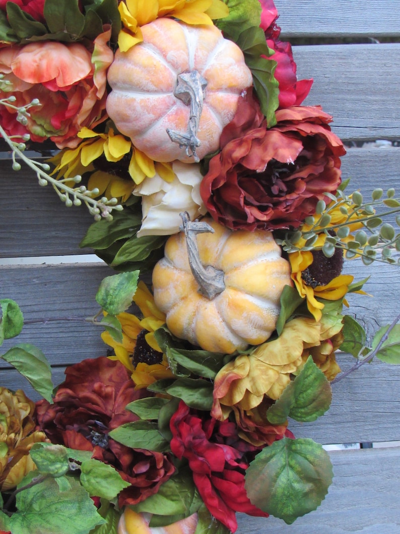 May include: A fall-themed wreath featuring red, orange, and yellow flowers, green leaves, and two faux pumpkins.