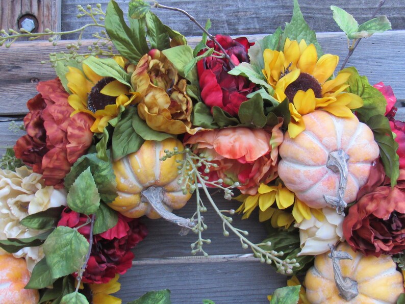 May include: A fall-themed wreath featuring sunflowers, peonies, and pumpkins. The wreath is decorated with greenery and has a rustic feel.