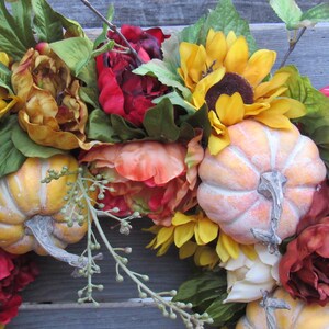 May include: A fall-themed wreath featuring red, orange, and yellow flowers, sunflowers, and small pumpkins. The wreath is crafted from artificial flowers and pumpkins.