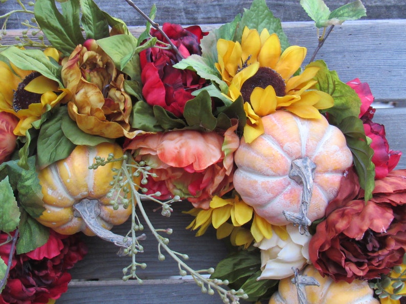 May include: A fall-themed wreath featuring red, orange, and yellow flowers, sunflowers, and small pumpkins. The wreath is crafted from artificial flowers and pumpkins.