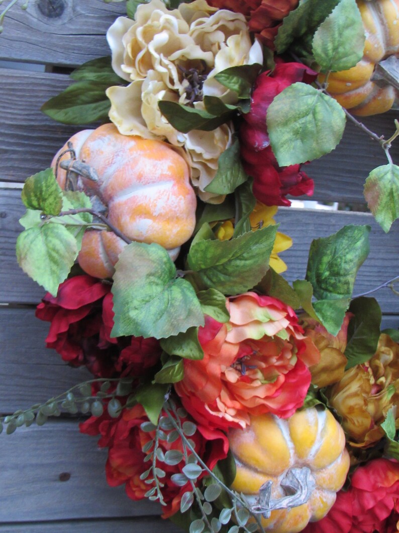 May include: A fall-themed wreath featuring orange and yellow pumpkins, red and orange peonies, and green leaves.